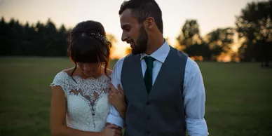 A bride and groom laughing together in the field as the sun sets behind the treeline